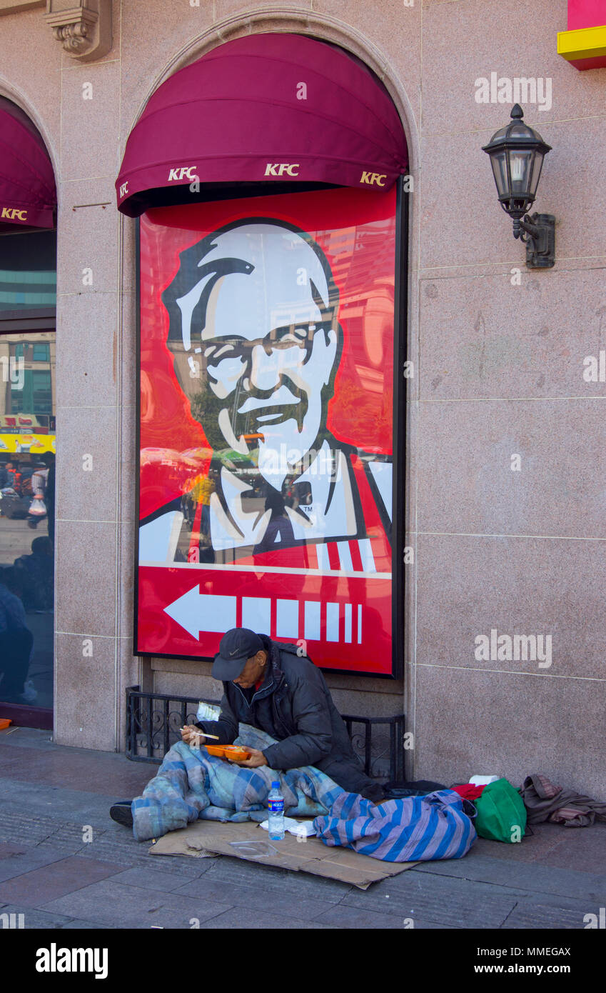 Ein Obdachloser auf dem Bürgersteig vor einem KFC-Restaurant in der Nähe von Beijing Railway Station in Peking, China, Sitzen, Essen zum Mitnehmen Essen mit Stäbchen. Stockfoto