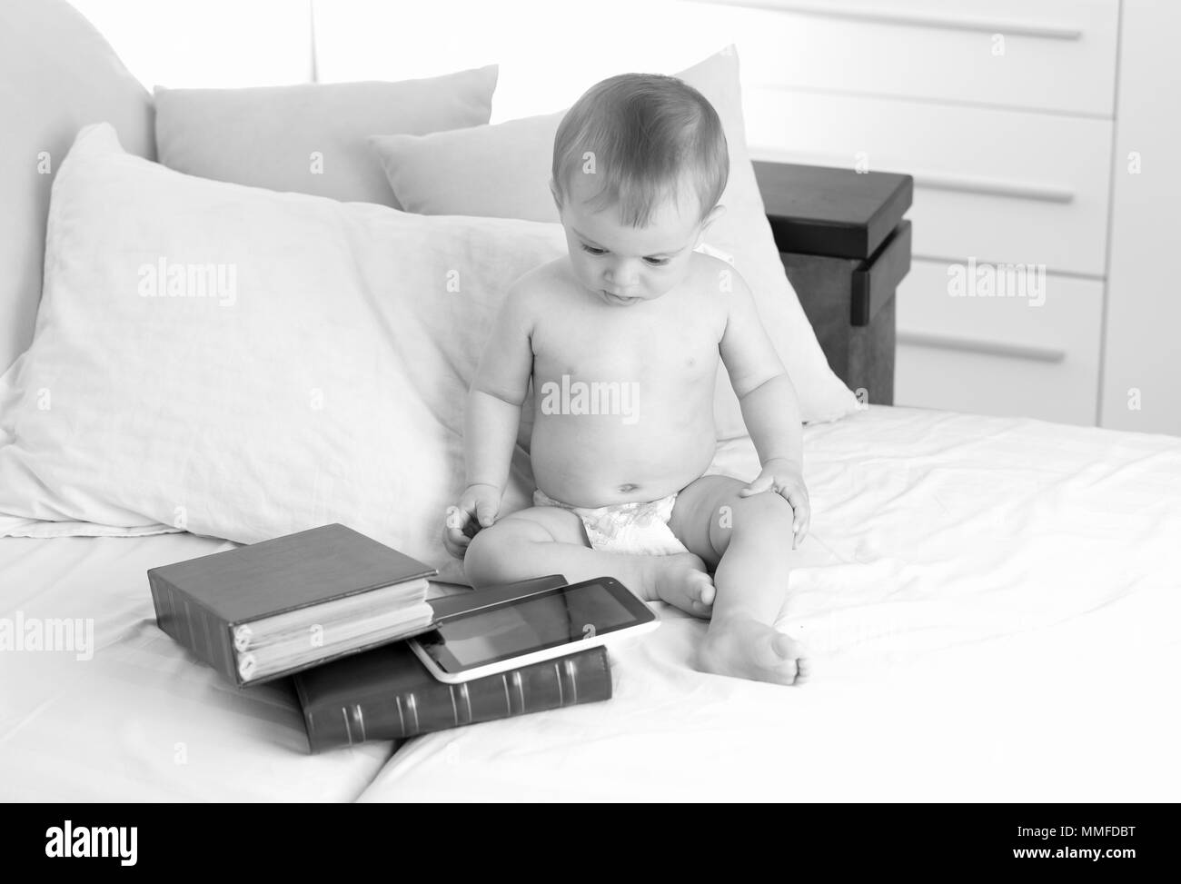 Schwarz-weiß Bild von adorable Baby in Windeln sitzen auf dem Bett mit großen atck von Büchern und digitalen Tablet. Konzept des smart kids und frühe Bildung mit dem Einsatz moderner Technologien und Computer Stockfoto