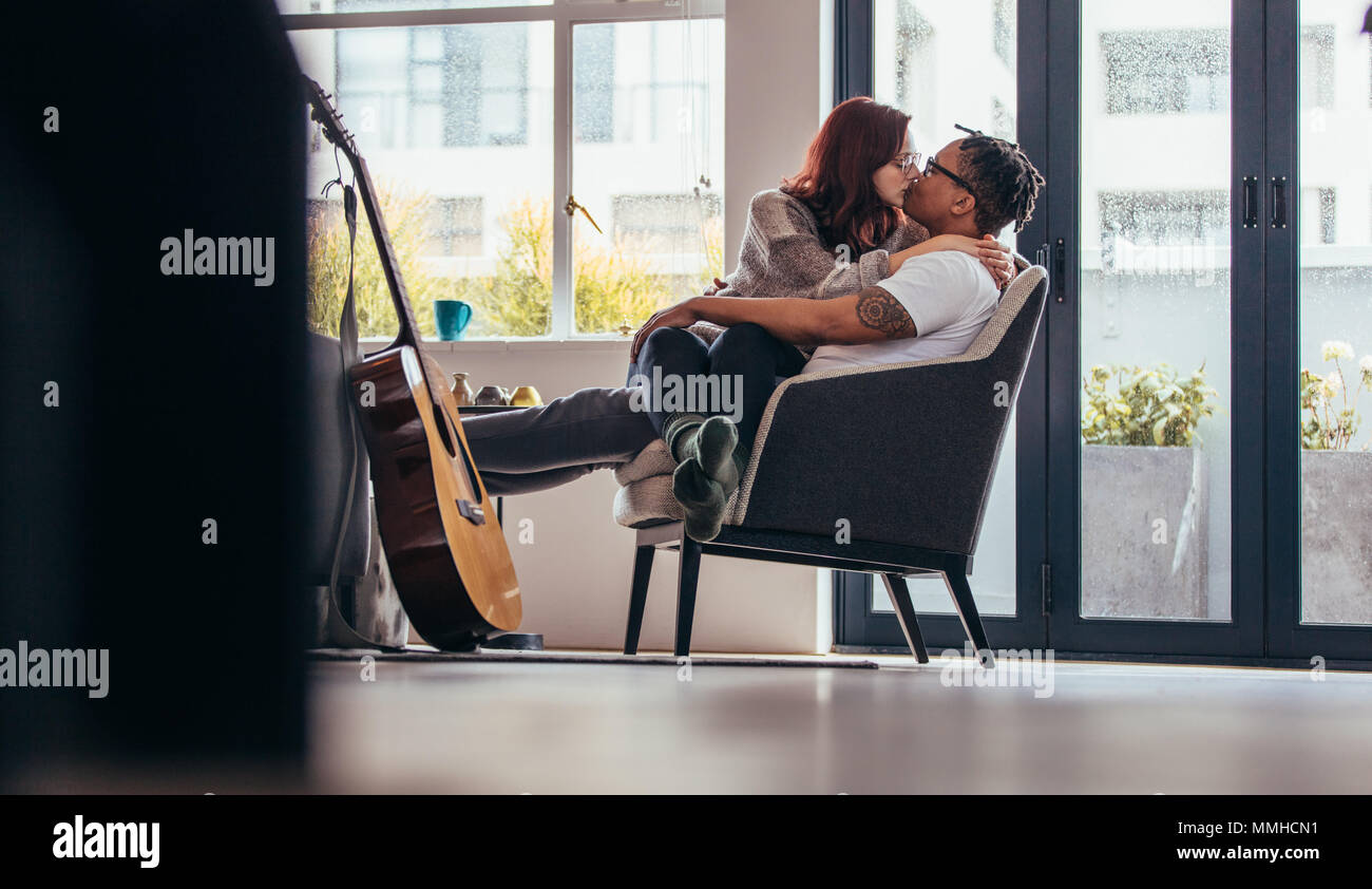 Interracial Paare auf einem Sessel und küssen einander sitzen. Frau sitzt auf dem Schoß der Mann und Küssen. Stockfoto