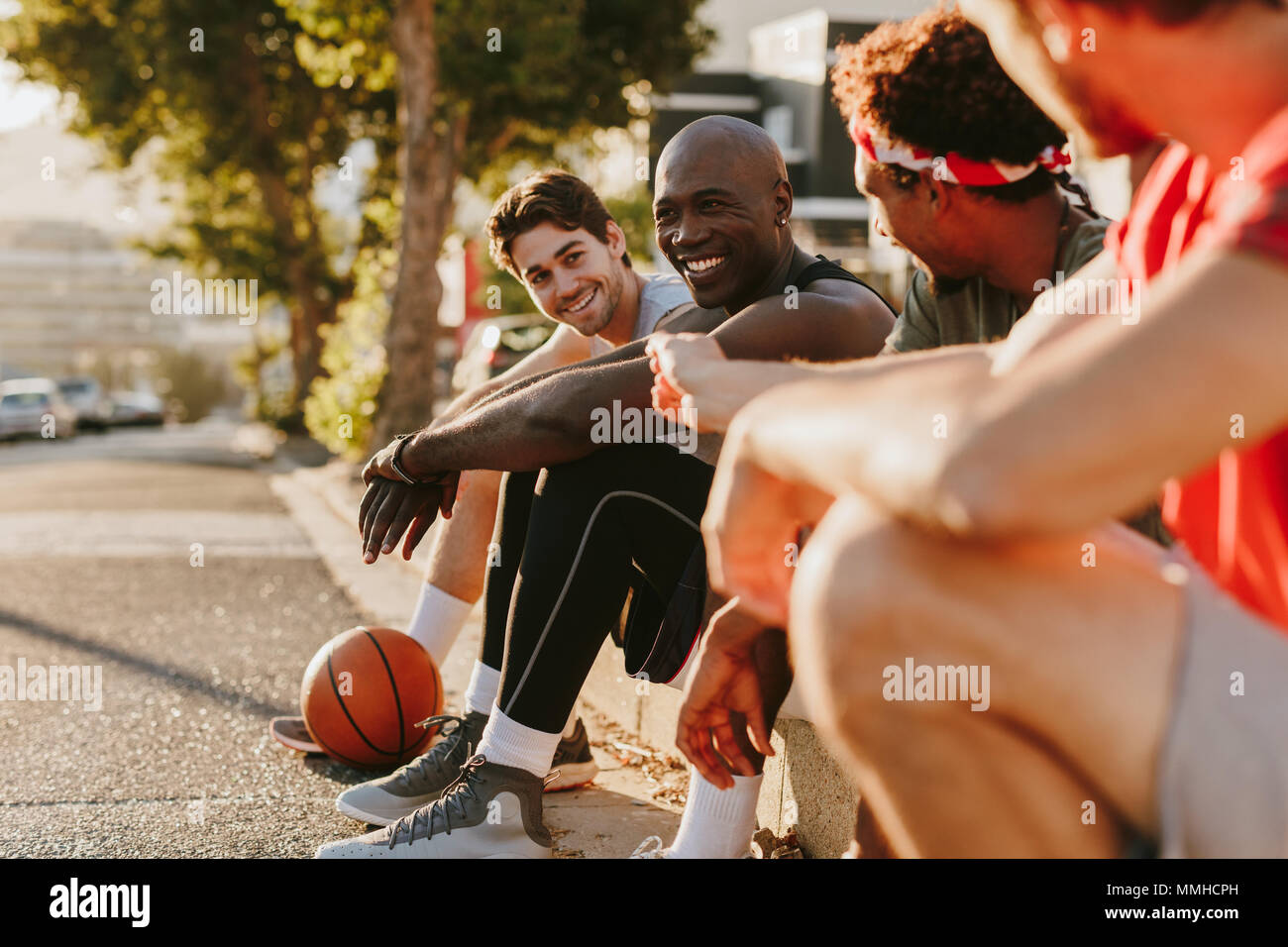Vier Basketball Spieler entspannen nach einem Spiel der Basketball sitzen auf Pflaster. Fröhliche Sportler sitzen auf Bürgersteig und diskutieren. Stockfoto