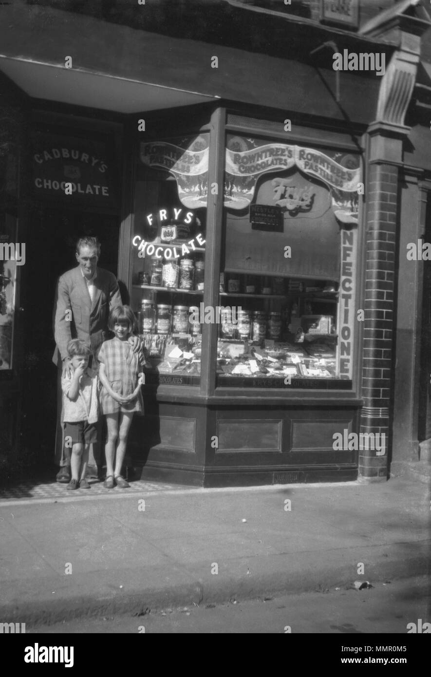 1930er Jahre, Vater mit seinen beiden Kindern am Eingang zu einem traditionellen Sweet Shop, England, UK. Cadbury's Schokolade geschrieben steht über dem Eingang und die Namen der anderen großen Süsswarenmarken des Tages, einschließlich Fry's, "Swiss made"-Nestle und Rowntree's gesehen werden kann. Stockfoto