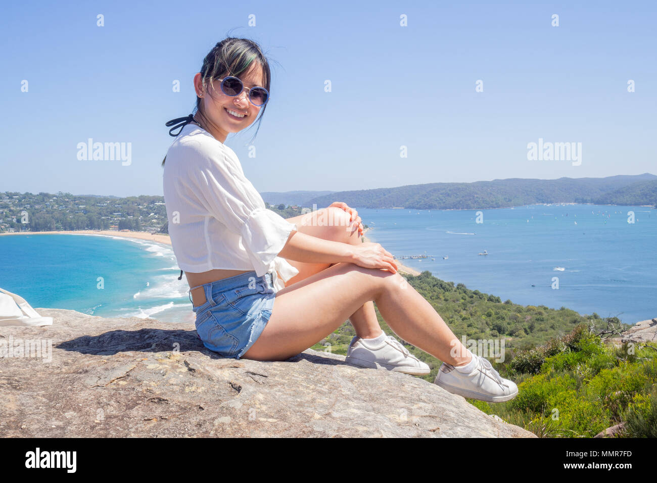 Junge Frau mit Sonnenbrille sitzt auf dem Hintergrund der Palm Beach auf einem Felsen am Leuchtturm in Barrenjoey, Sydney, Australien. Stockfoto