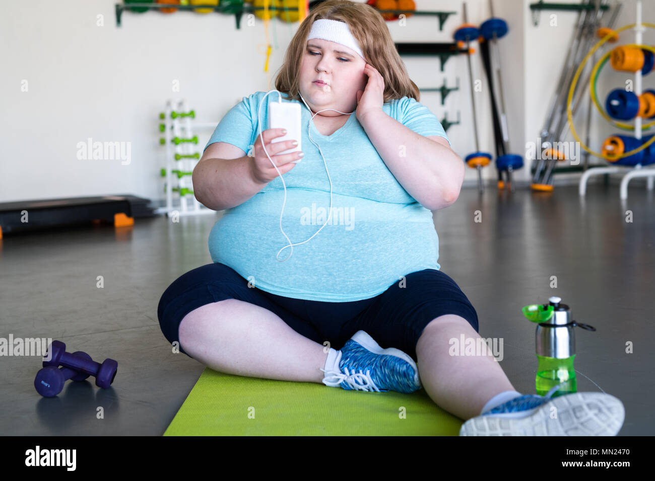 In voller Länge Porträt der übergewichtige junge Frau sitzt auf Yoga Matte auf dem Boden und Hören von Musik bereit Fitnesstraining zu starten Stockfoto