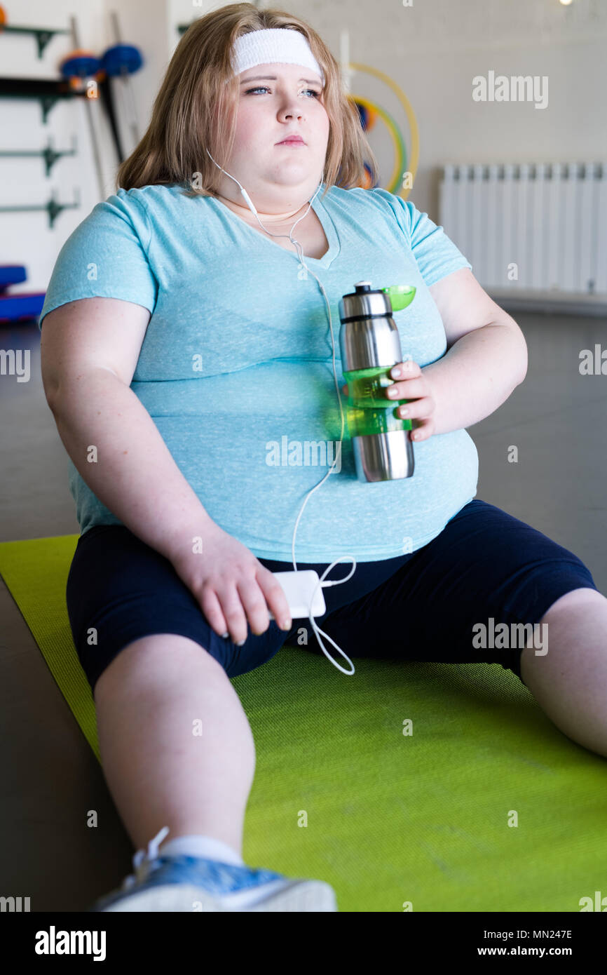 Volle Länge Portrait von Müde beleibte Frau sitzt auf Yoga Matte auf dem Boden und Hören von Musik entspannen nach Fitness Workout Stockfoto