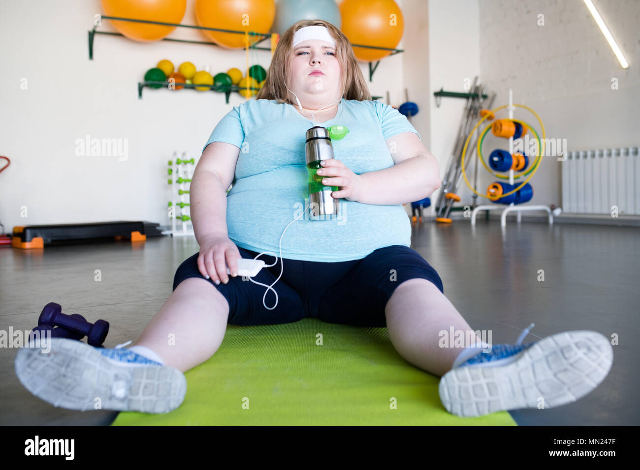Volle Länge Portrait von Müde beleibte Frau sitzt auf Yoga Matte auf dem Boden und Hören von Musik ausruhen nach Fitness Workout Stockfoto