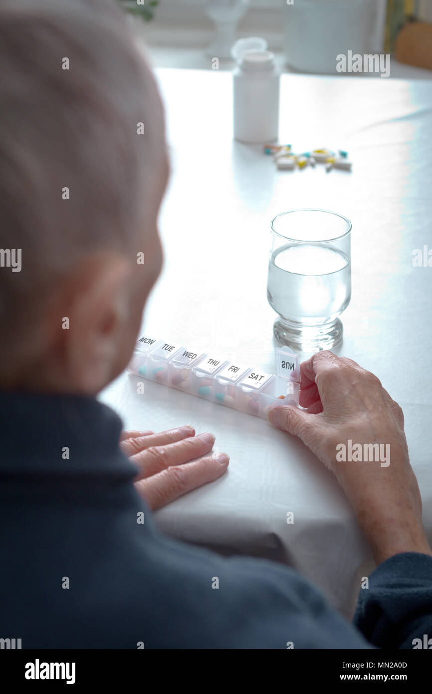 Alte Frau an einem Tisch zu Hause sitzen mit einem Glas Wasser und viele Kapseln in verschiedenen Farben in einer Vorsortierten wöchentliche Tablettenspender. Stockfoto