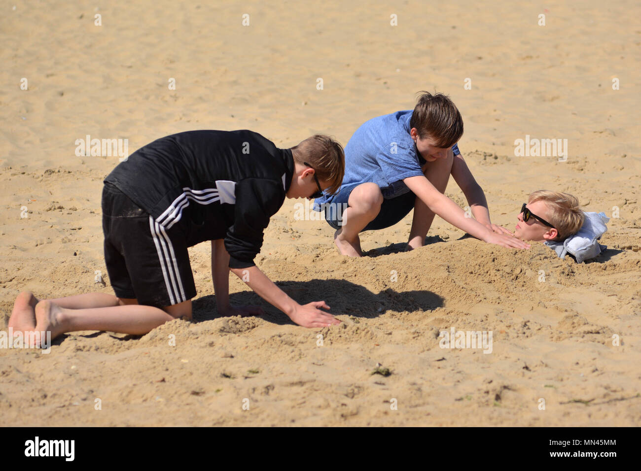 Zwei Jungen, die einen anderen Jungen vergraben im Sand am Strand an der Südküste Badeort Bournemouth, Dorset, Großbritannien, im warmen Sonnenschein. Stockfoto