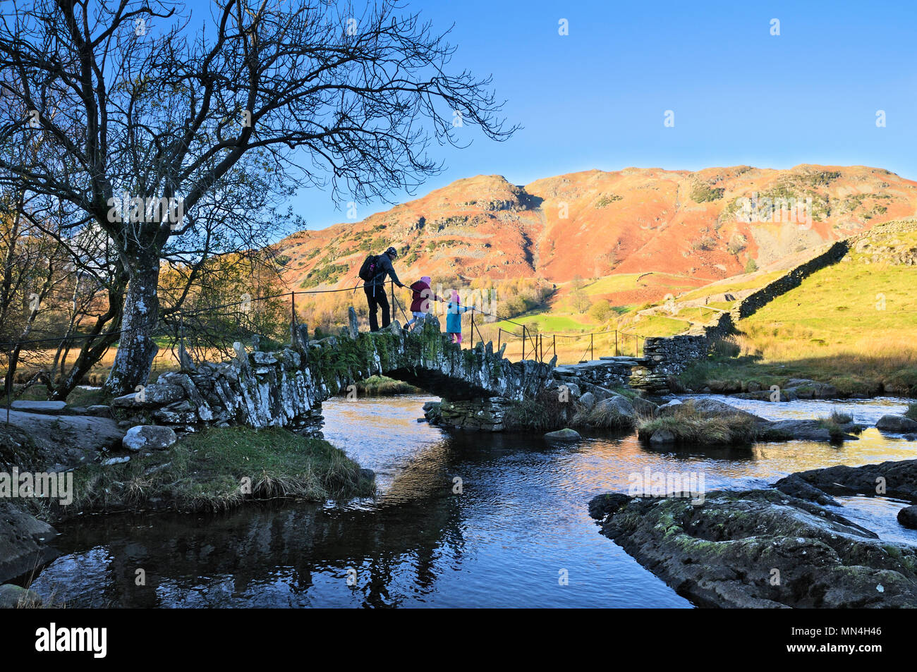 Vater und Kinder zu Fuß über Slater Brücke über den Fluss Brathay vor dem Hintergrund der Lingmoor fiel, Little Langdale, Lake District, Großbritannien Stockfoto