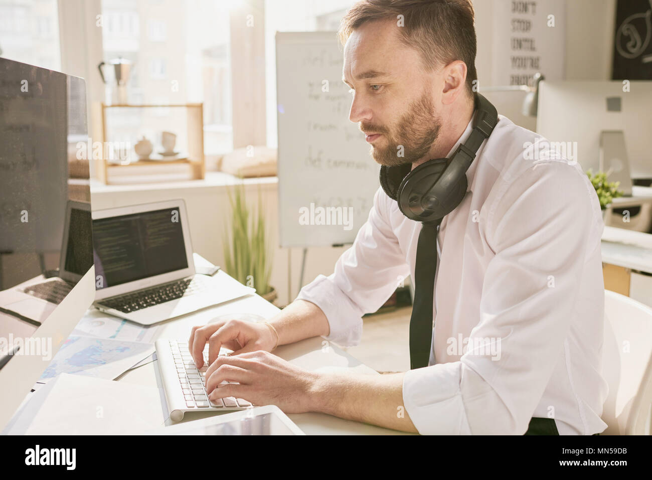 Seitenansicht Portrait von gut aussehenden bärtigen Geschäftsmann mit Hemd und Krawatte sitzt am Schreibtisch im Büro und den Einsatz moderner Computer, Eingabe, mit Kopfhörern Stockfoto