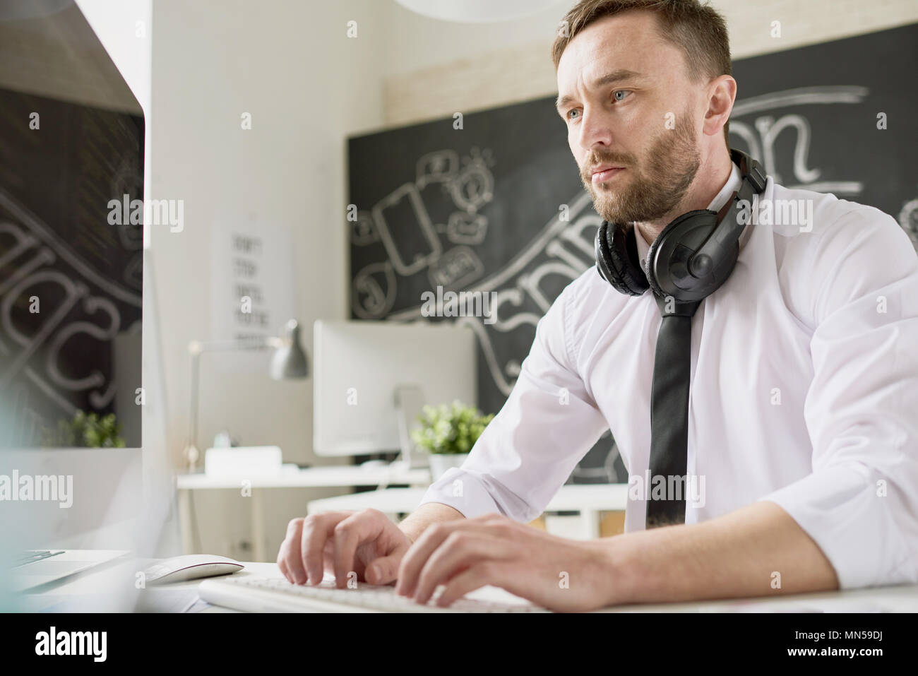 Portrait von gut aussehenden bärtigen Geschäftsmann mit Hemd und Krawatte sitzt am Schreibtisch im Büro und den Einsatz moderner Computer, Tippen, mit Kopfhörer an seinem Hals Stockfoto