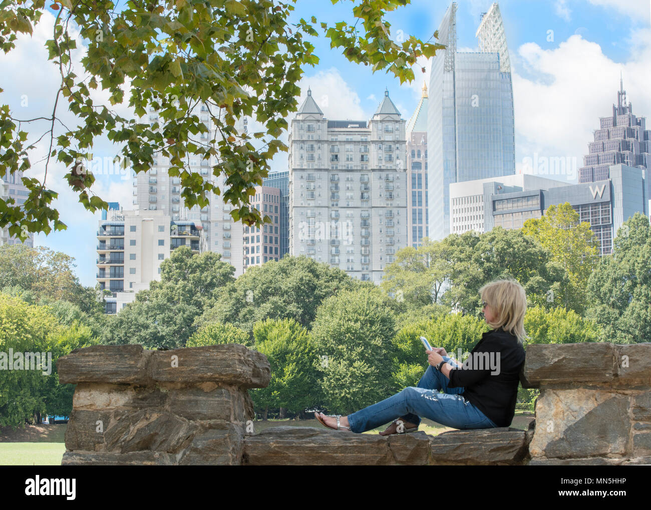 Schöne Frau sitzend auf der Steinmauer in Piedmont Park das Spielen mit Ihrem Handy. Atlanta Skyline im Hintergrund. Stockfoto