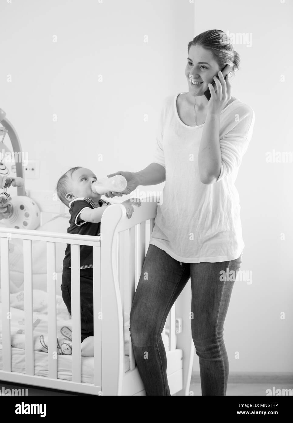 Schwarz-weiß Bild von jungen beschäftigte Frau im Gespräch per Telefon und geben Milch für Ihr Baby Sohn Stockfoto