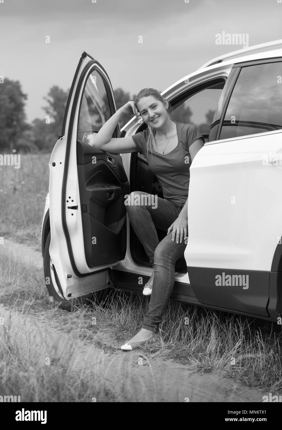 Schwarz-weiß Bild von lächelnden jungen Frau, die mit dem Auto anreisen, geöffnete Tür in der Kamera auf der Suche Stockfoto