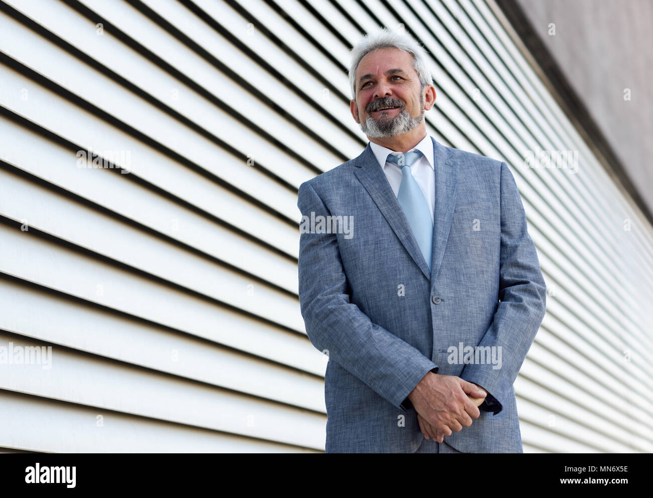 Portrait eines älteren Geschäftsmann mit Waffen außerhalb der modernen Bürogebäude gekreuzt. Erfolgreiche Mann mit Anzug und Krawatte im städtischen Hintergrund lächelnd. Stockfoto