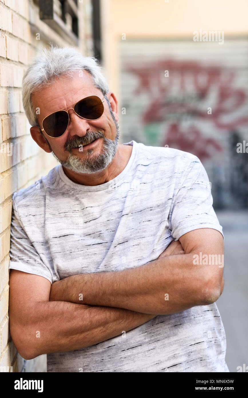 Reifen Mann an der Kamera im städtischen Hintergrund lächelnd. Älterer Mann mit weissem Haar und Bart legere Kleidung und Pilotenbrillen. Stockfoto