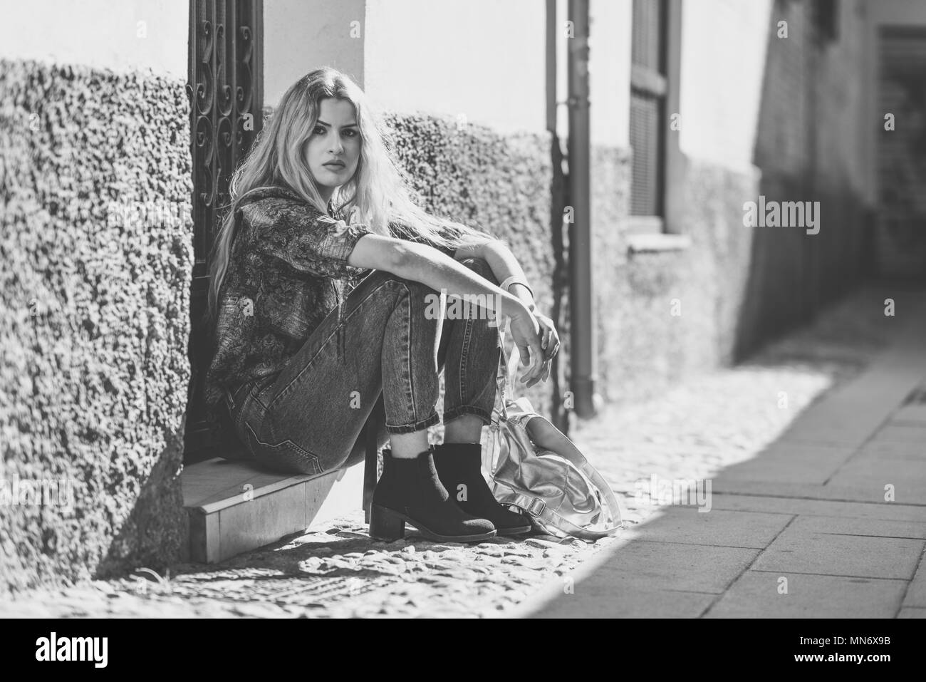 Ernsthafte blonde Frau, Modell der Mode, sitzend auf dem Boden im städtischen Hintergrund. Schöne Mädchen mit T-Shirt und eine blaue Jeans. Stockfoto