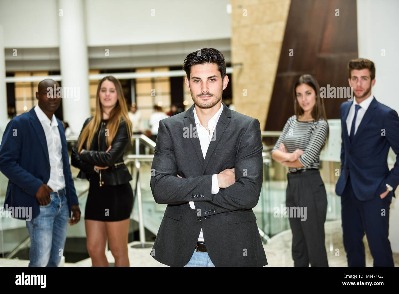 Junge Unternehmer Führer an Kamera suchen mit Waffen im Bürogebäude gekreuzt. Gruppe von muti-ethnischen Menschen im Hintergrund Stockfoto