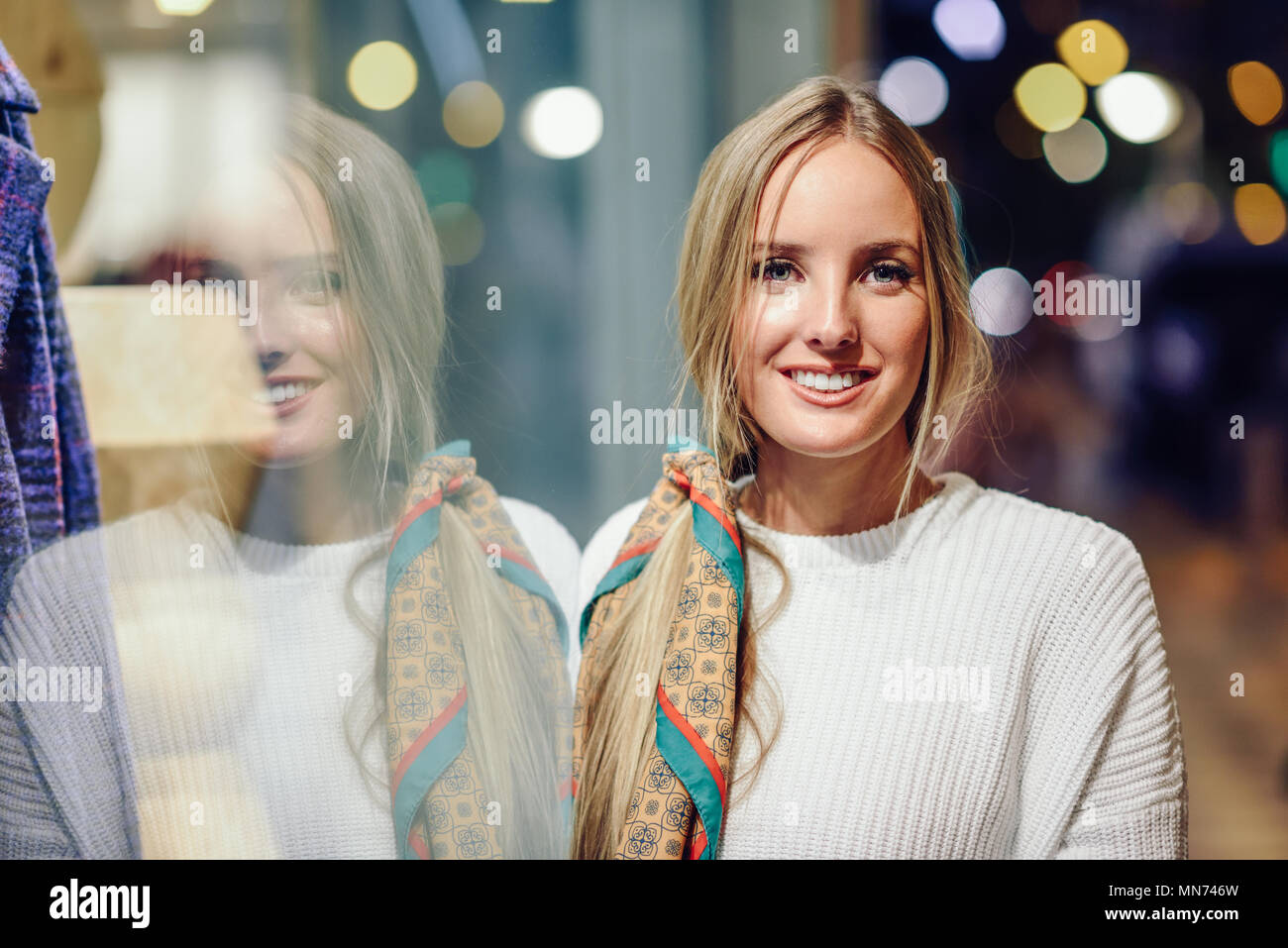 Blonde Mädchen tragen weiße Pullover in der Straße lächelnd mit Defokussierten Lichter der Stadt im Hintergrund. Hübsche Frau mit Zopf Frisur in der Nacht in einem Schaufenster wider. Stockfoto