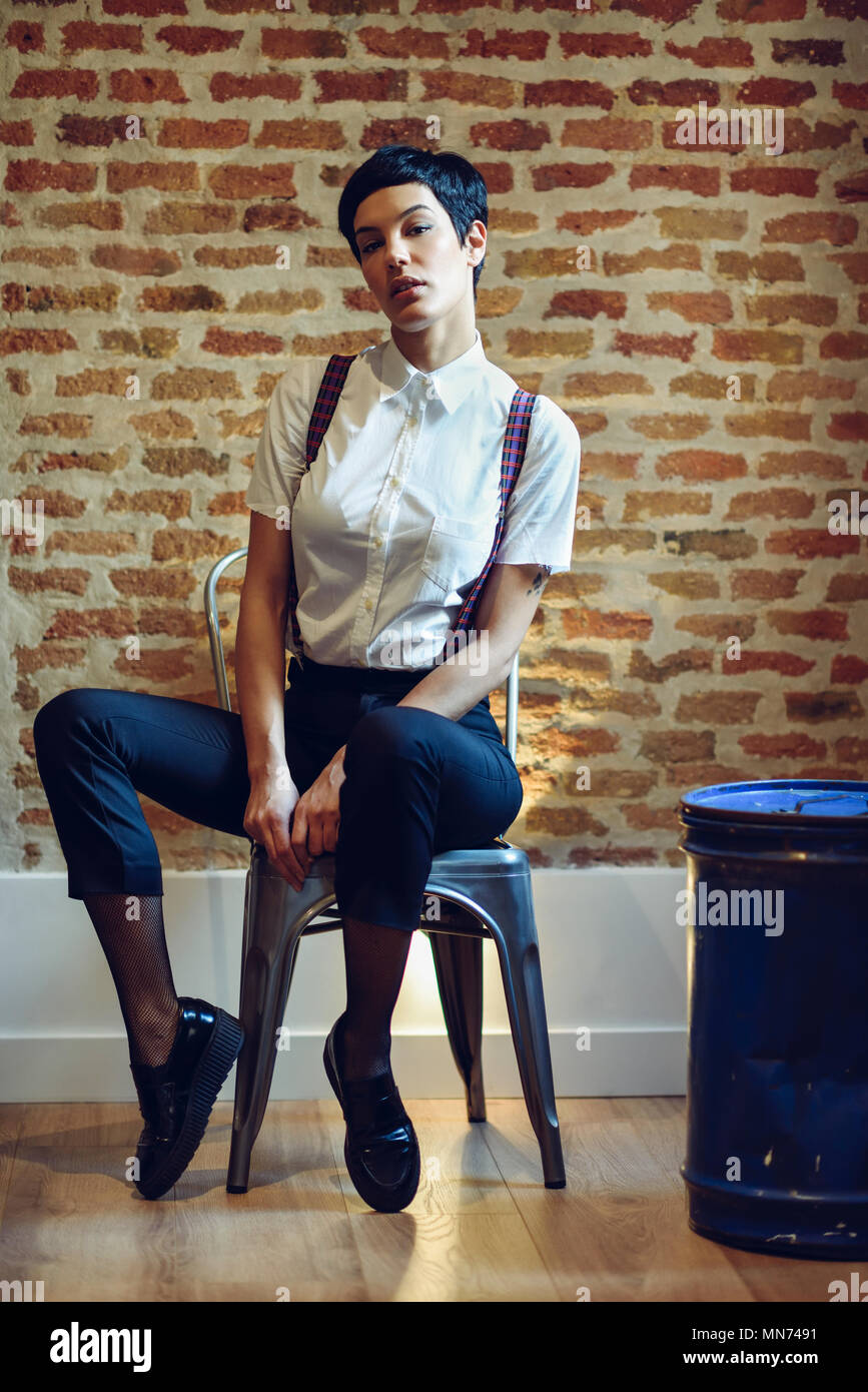 Schöne junge Frau, Modell der Mode, mit sehr kurzen Haarschnitt, an der Kamera sitzt auf Stuhl suchen, mit einer Mauer im Hintergrund. Schönheit und Frisuren Konzept. Stockfoto