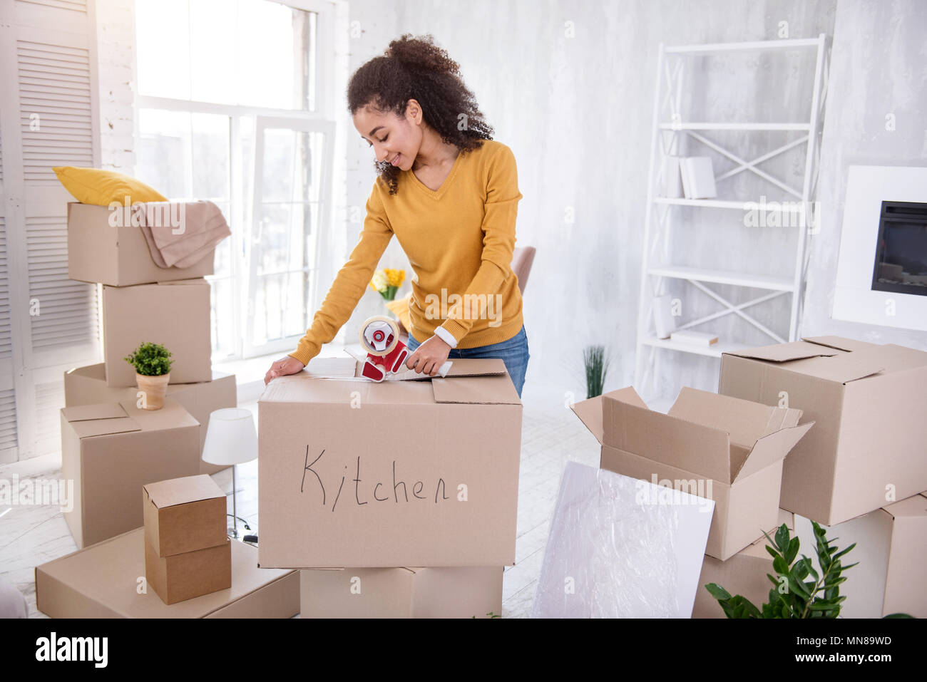 Fröhliche lockigen dunkelhaarigen Mädchen taping Box mit Küchengeräte Stockfoto