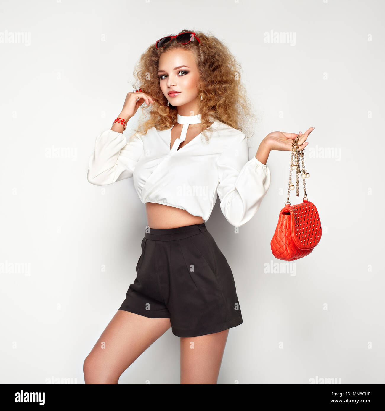 Blonde Frau im Sommer Bluse und Hose. Girl posiert auf weißem Hintergrund. Rote Handtasche. Stilvolle curly Frisur. Glamour Dame in stilvolle Sonnenbrillen Stockfoto