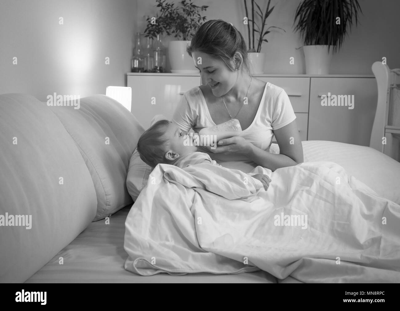 Schwarz-weiß Foto der jungen Mutter stillen ihr Baby Sohn mit Milch aus der Flasche in der Nacht Stockfoto