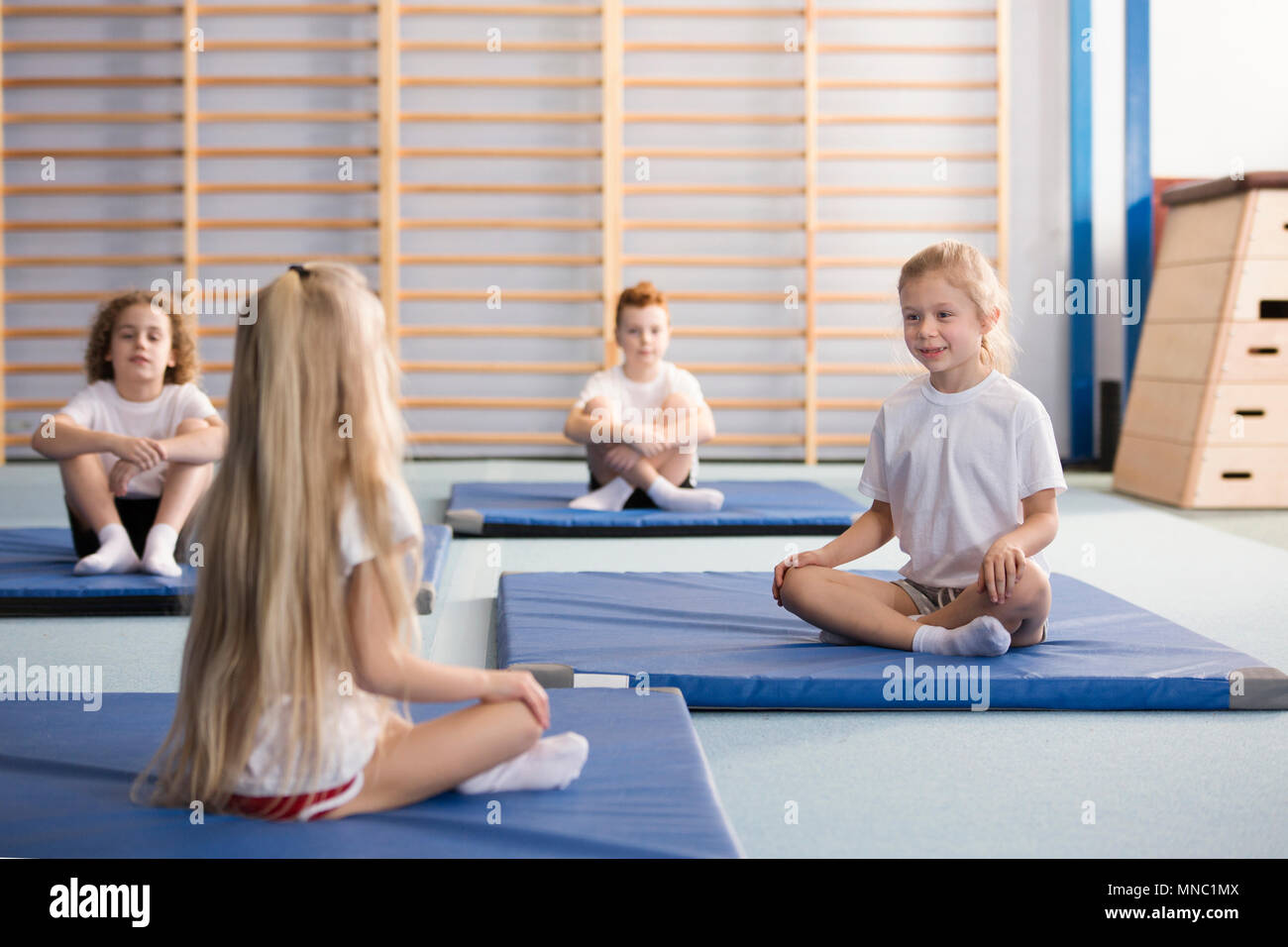 Glückliche junge Mädchen sitzen einander gegenüber auf blauen Matten Trainieren mit ihren Beinen während PE-Klassen in der Grundschule Turnhalle Innenraum gekreuzt Stockfoto