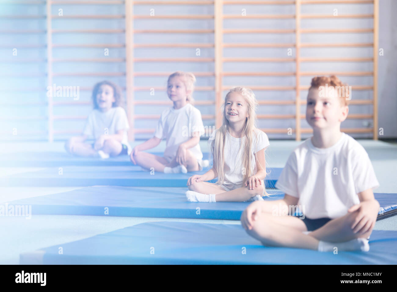 Neugierig und erstaunt, die Kinder in der Schule Sportswear sitzen in einer Turnhalle Innenraum während der außerschulischen Yoga class und bis in die Richtung einer flar suchen Stockfoto