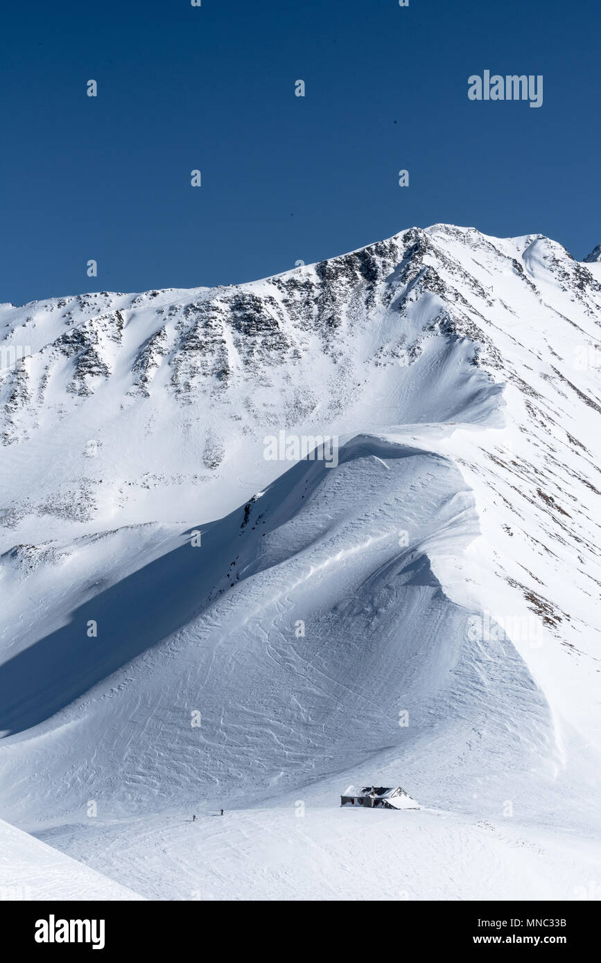Zwei Skifahrer verlassen Berghütte teilweise durch Schnee an der Unterseite der Bergrücken und Gipfel über bluebird Tag bedeckt im Winter Stockfoto