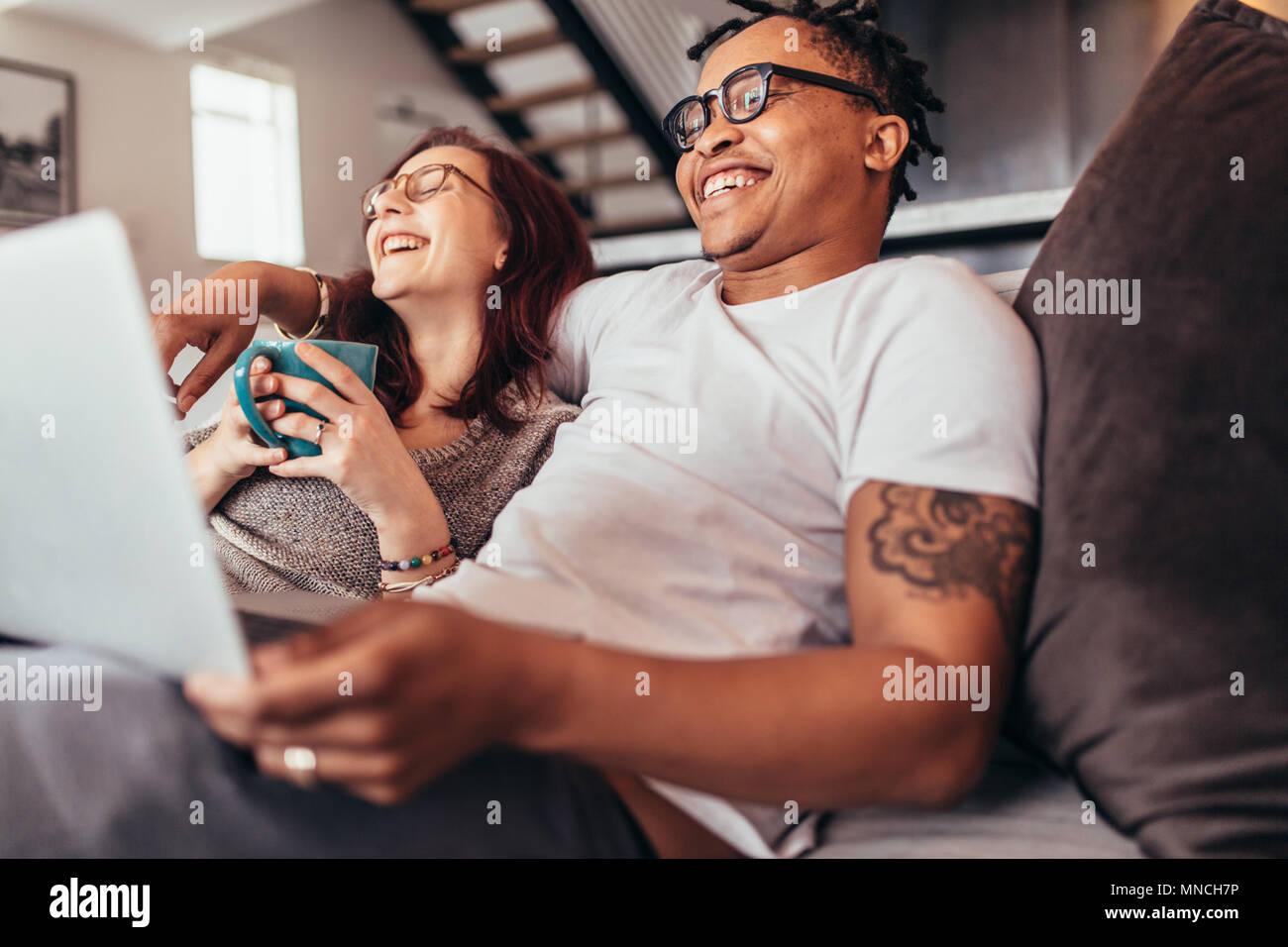 Fröhliche multi-ethnischen Paar zusammen entspannt auf einem Sofa mit Laptop und eine Tasse Kaffee. Lächelnd Mann und Frau auf der Couch zu Hause sitzen. Stockfoto