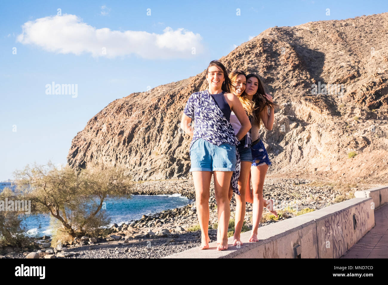 Drei junge Frauen, die Freunde gehen gemeinsam an einer Wand in der Nähe der Strand auf Teneriffa. Die Ferienhäuser unter einem blauen Himmel mit gelben Sonne genießen. Hände von den Händen. Stockfoto Drei junge Frauen, die Freunde gehen gemeinsam an einer Wand in der Nähe der Strand auf Teneriffa. Die Ferienhäuser unter einem blauen Himmel mit gelben Sonne genießen. Hände von den Händen. Stockfoto