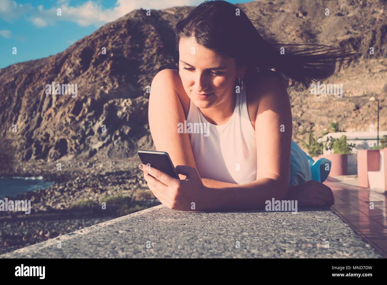 Schöne lange Haare spanische Frau Mobil Telefon Nachricht bei einem Urlaub in Teneriffa zu senden. Legen Sie auf einer Wand in der Nähe des Strandes unter einem blauen Himmel w Stockfoto