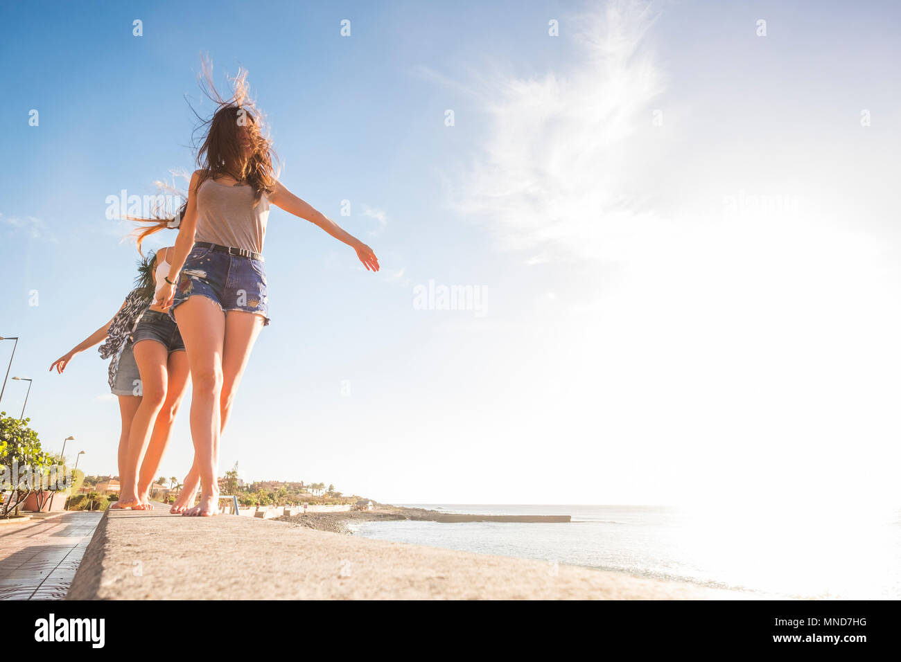 Drei junge Frauen, die Freunde gehen gemeinsam an einer Wand in der Nähe der Strand auf Teneriffa. Die Ferienhäuser unter einem blauen Himmel mit gelben Sonne genießen. Hände von den Händen. Stockfoto Drei junge Frauen, die Freunde gehen gemeinsam an einer Wand in der Nähe der Strand auf Teneriffa. Die Ferienhäuser unter einem blauen Himmel mit gelben Sonne genießen. Hände von den Händen. Stockfoto