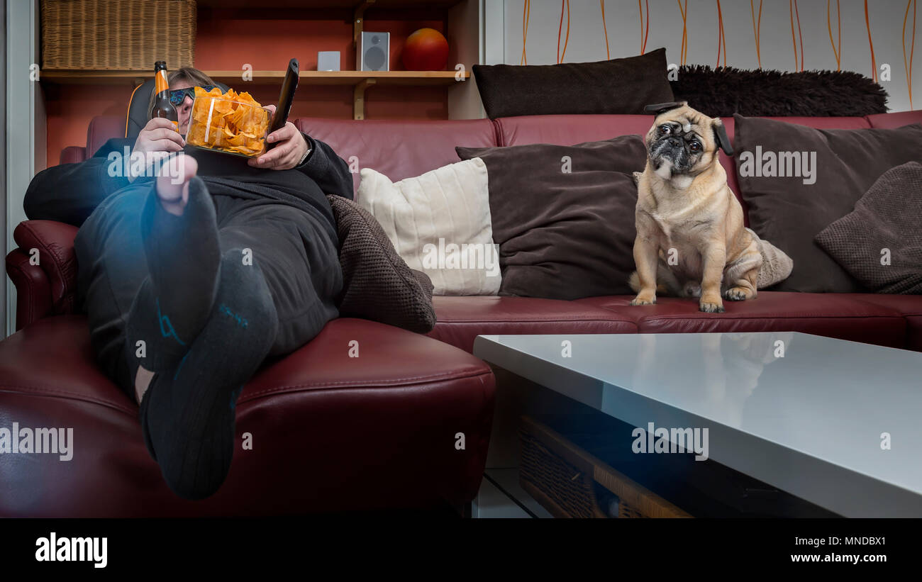 Ein Mensch isst Chips vor dem Fernseher. Sein Hund sitzt neben ihm auf der Couch. Der Mann hat ein Loch in die Strümpfe und eine 3D-Brille auf. Frosch p Stockfoto