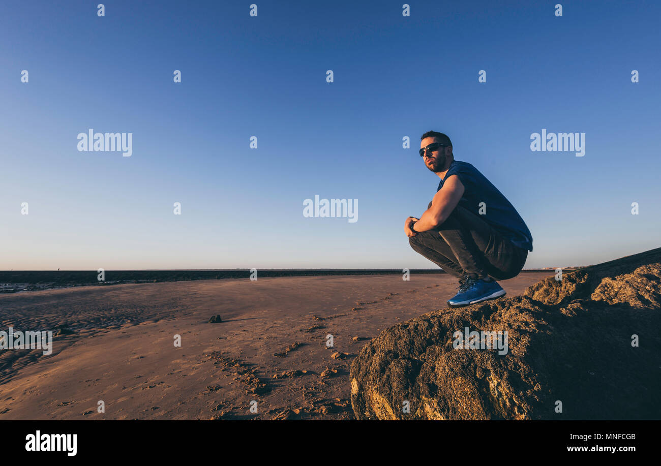Junger Mann kauert auf einem Felsen mit Sonnenbrille am Strand von Sanlucar de Barrameda Stockfoto