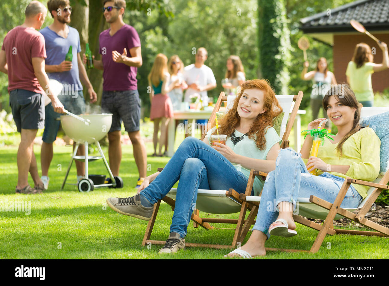 Zwei Mädchen sitzen auf Liegestühlen mit Getränken, Männer Grillen Stockfoto