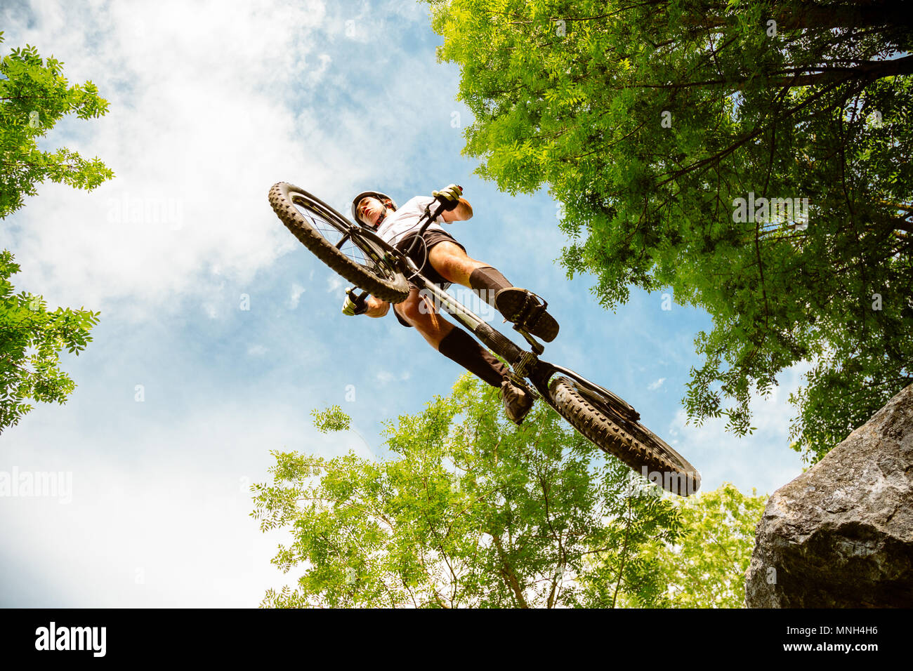 Junge Radfahrer fliegen mit seinem Fahrrad von einem Felsen im Wald. Extreme Low Angle View Stockfoto