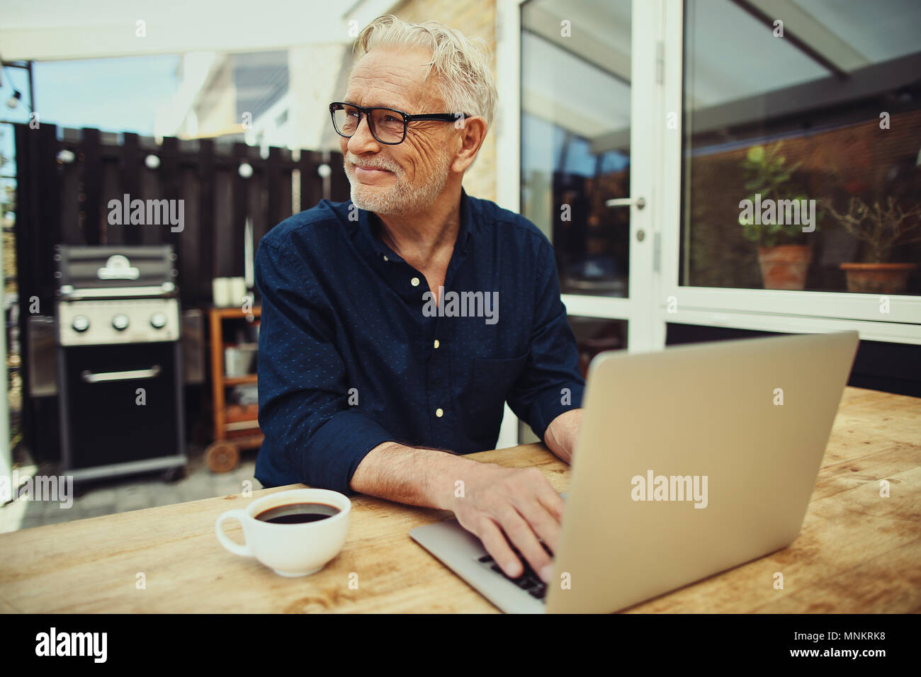 Lächelnd älterer Mann, trinken eine Tasse Kaffee und online arbeiten mit einem Notebook während an einem Tisch draußen auf der Terrasse sitzen Stockfoto