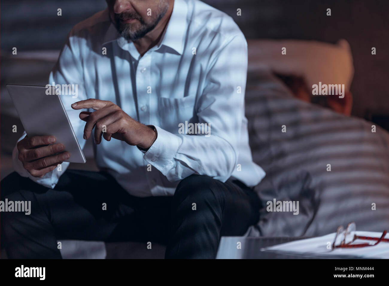 Low Angle View reifer Mann sitzt auf einem Bett in einem Zimmer und halten eine Tablette in der Hand mit einer Frau im Bett schlafen - Abdeckung in einer unscharfen backgro Stockfoto