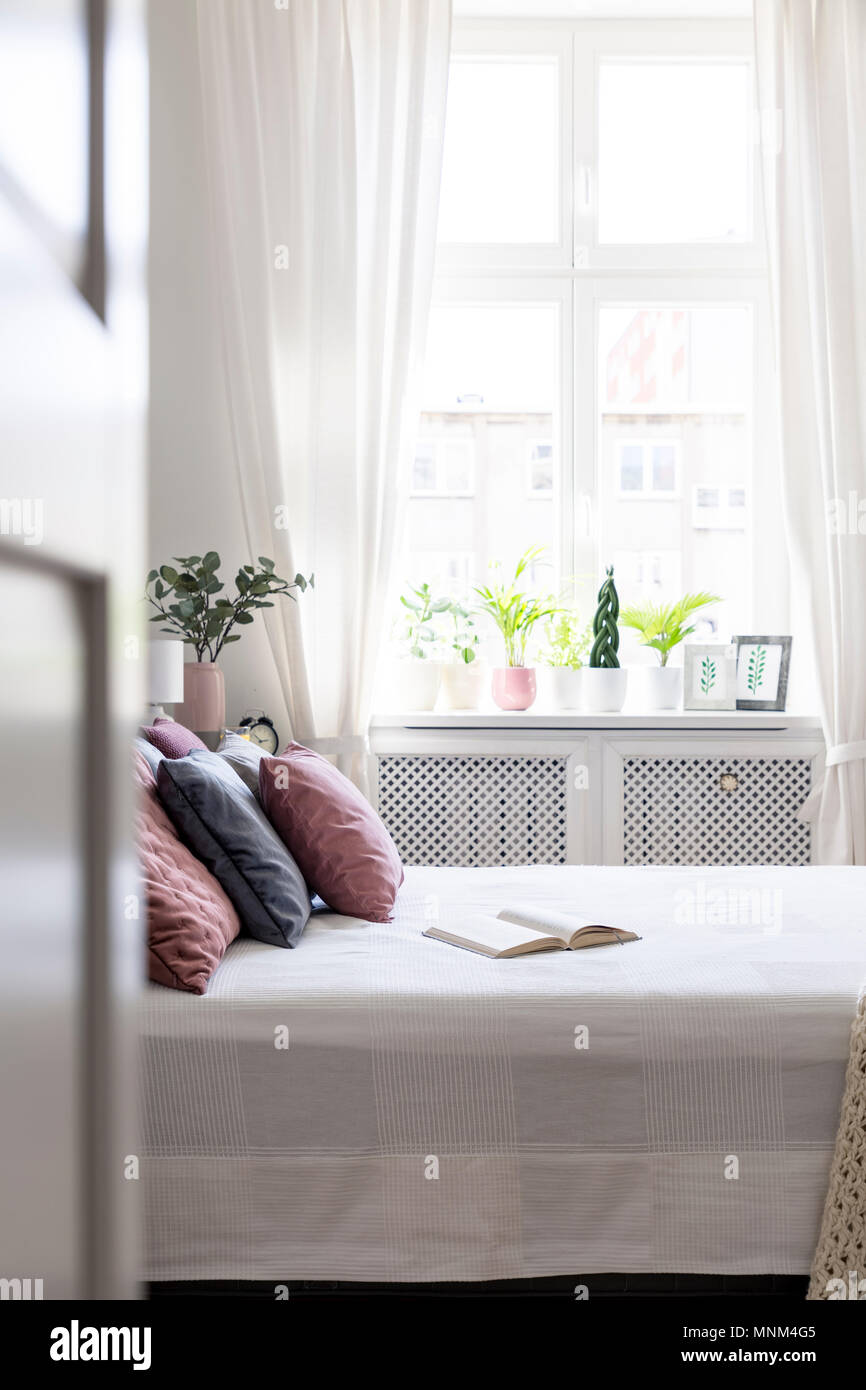 Buch und rosa Kissen auf dem Bett in weissen Schlafzimmer Innenraum mit Fenster. Real Photo Stockfoto