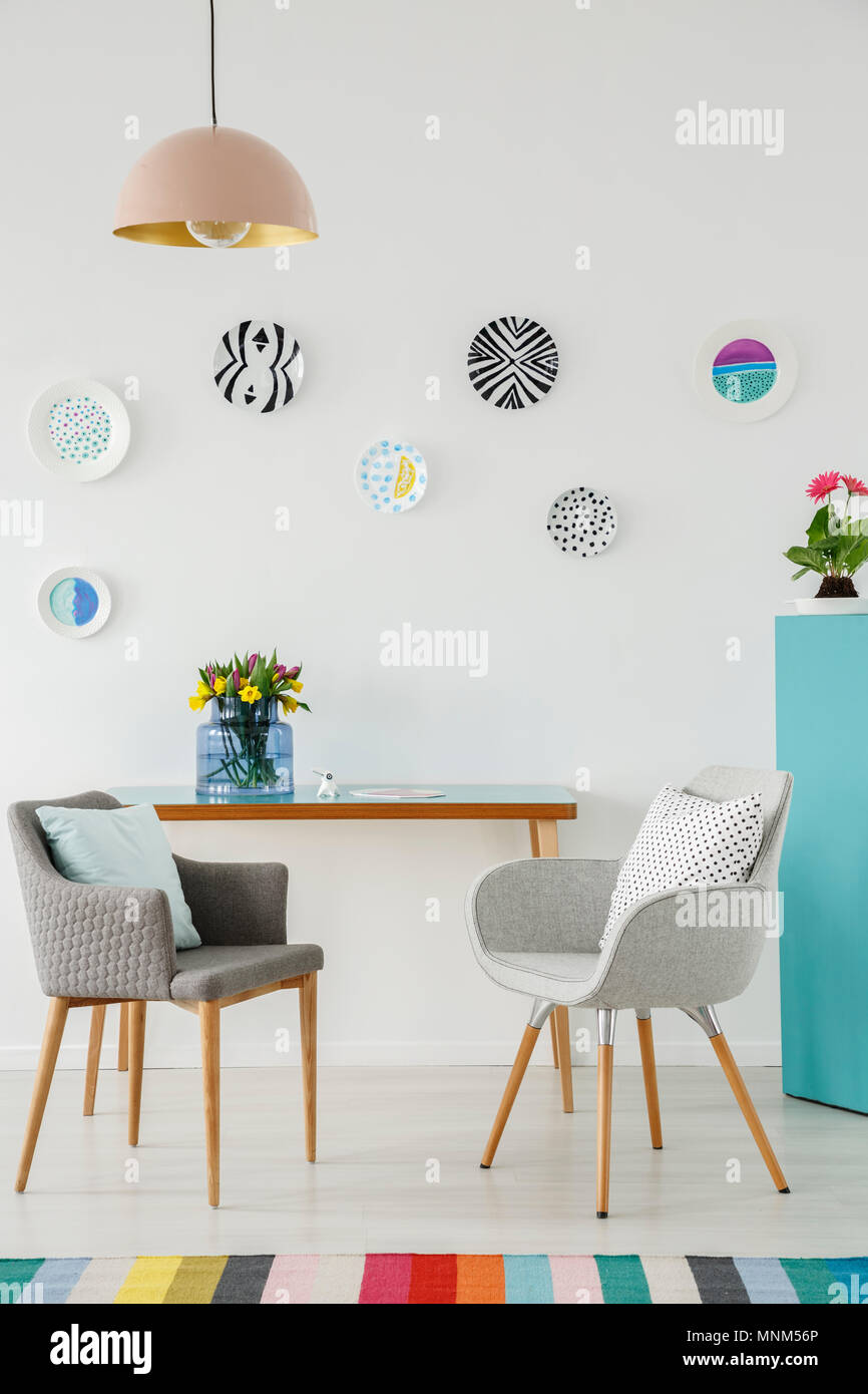 Kreative Wohnzimmer Interieur mit Platten an der Wand, auf dem Tisch, grauen Sesseln, Blumen und bunten Teppich Stockfoto