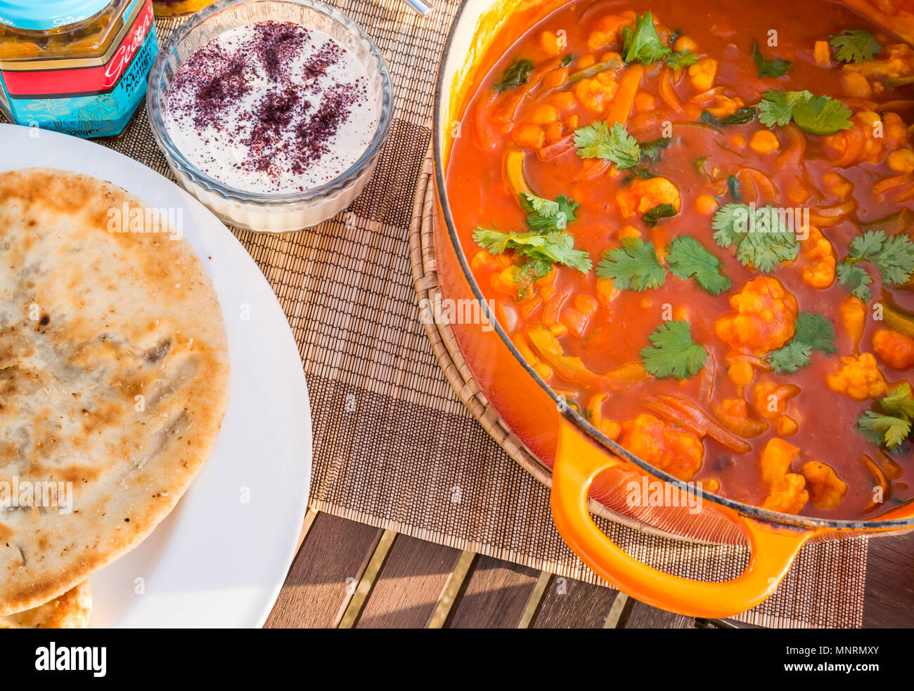 In der Nähe von großen orange Le Creuset Topf Topf auf sonnenbeschienenen Terrasse Tisch mit Blumenkohl Gemüse Curry, mit Sumach und Naan Brot Joghurt Stockfoto