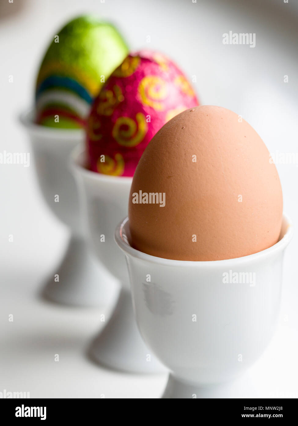 Schokolade Ostereier und eine braune Henne Ei im Eierbecher: Eine Reihe von drei Eier in drei weiße Porzellan Eierbecher auf einem weißen Hintergrund. Stockfoto