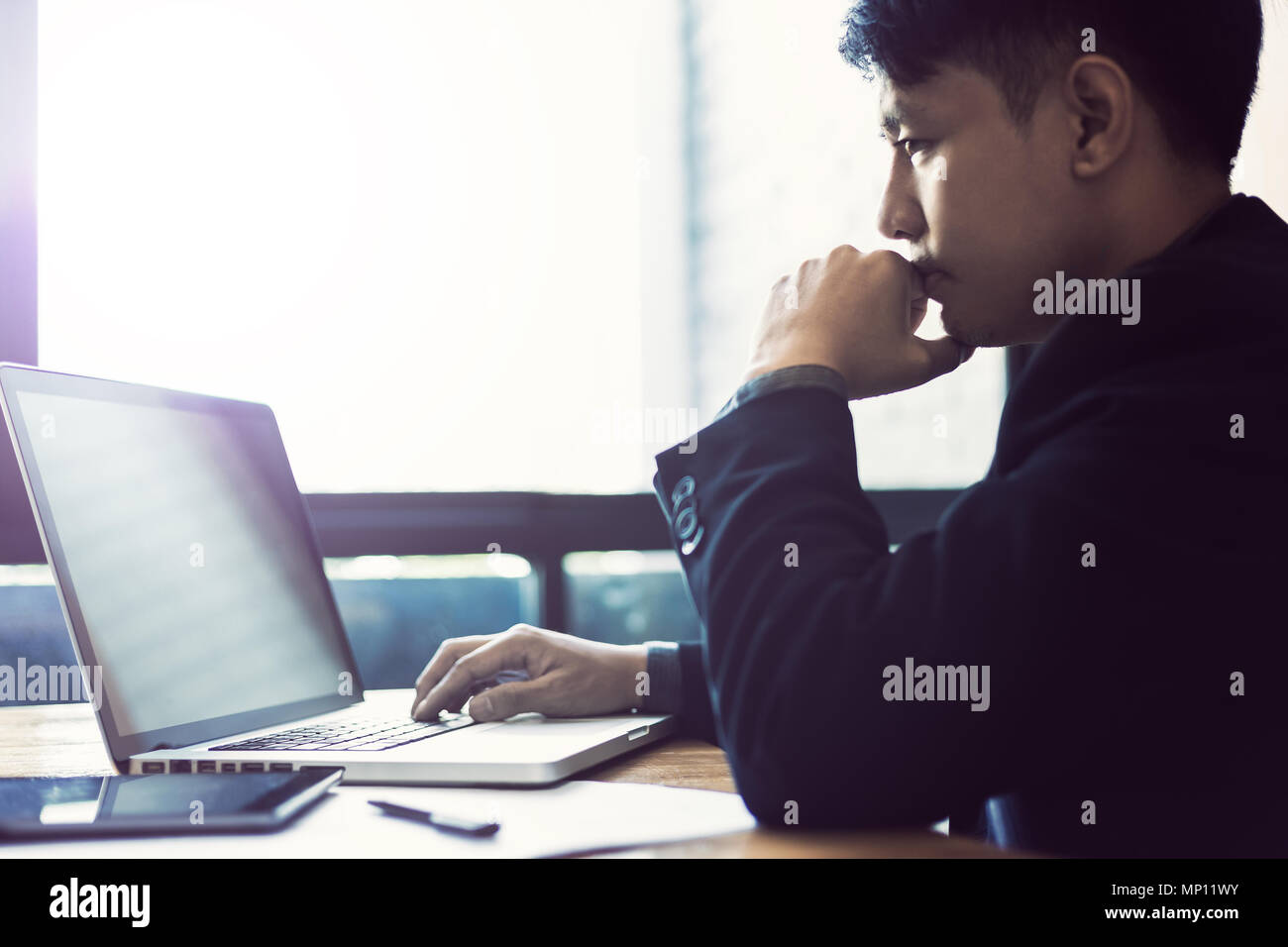 Junge asiatische Geschäftsmann mit der Arbeit an seinem Computer in seinem Büro in der Nähe von große, helle Fenster, selektive Schärfe und Unschärfe Farbe, Tonisierung, gut für Business Theme Stockfoto