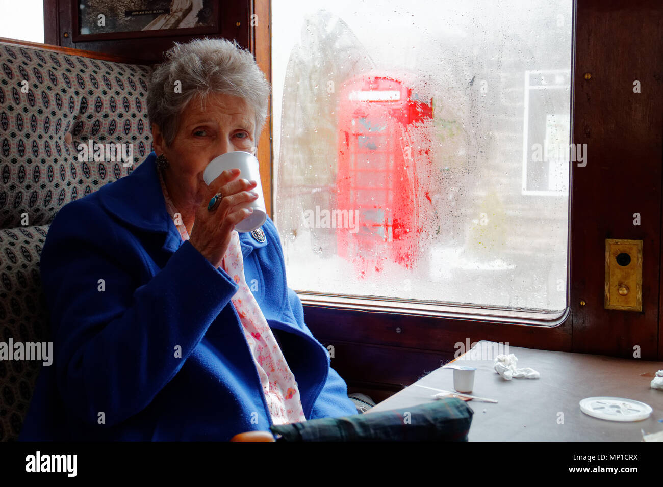 Eine ältere Frau trinkt Tee auf der Peak Bahn Dampfzug an einem regnerischen Tag in in Bakewell, Derbyshire mit einem roten Feld außerhalb Stockfoto