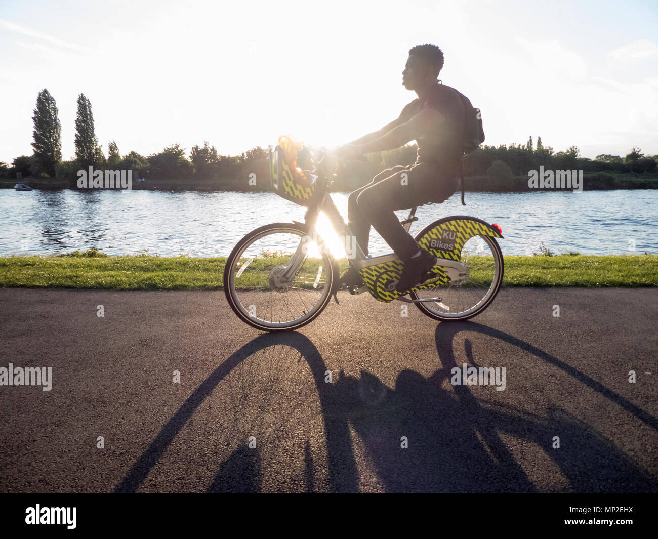 Hintergrundbeleuchteter junger Mann, der auf Der Themse auf dem Kingston University e-Bike mit langem Schatten radeln kann Stockfoto
