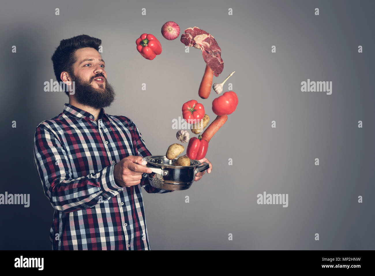 Kochen man Concept, lächelnd bärtigen Mann in kariertes Hemd, Nach oben Drop Fleisch und Gemüse aus der Pfanne, Studio shot auf grauem Hintergrund Stockfoto