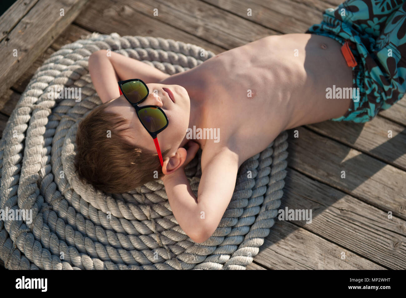 Niedliche Kind in der Sonnenbrille liegen auf hölzernen Pier mit Seil unter der heißen Sommersonne Stockfoto