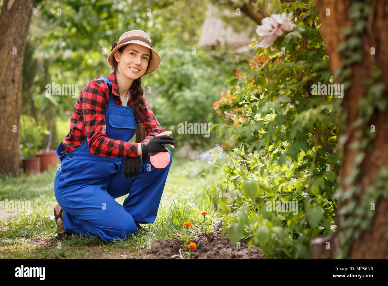 Porträt der glückliche junge weibliche Gärtner in Arbeitskleidung holding Sprühflasche mit grünen verschwommen Hof im Hintergrund. Gartenbau und Pflege der Pflanzen ein Stockfoto