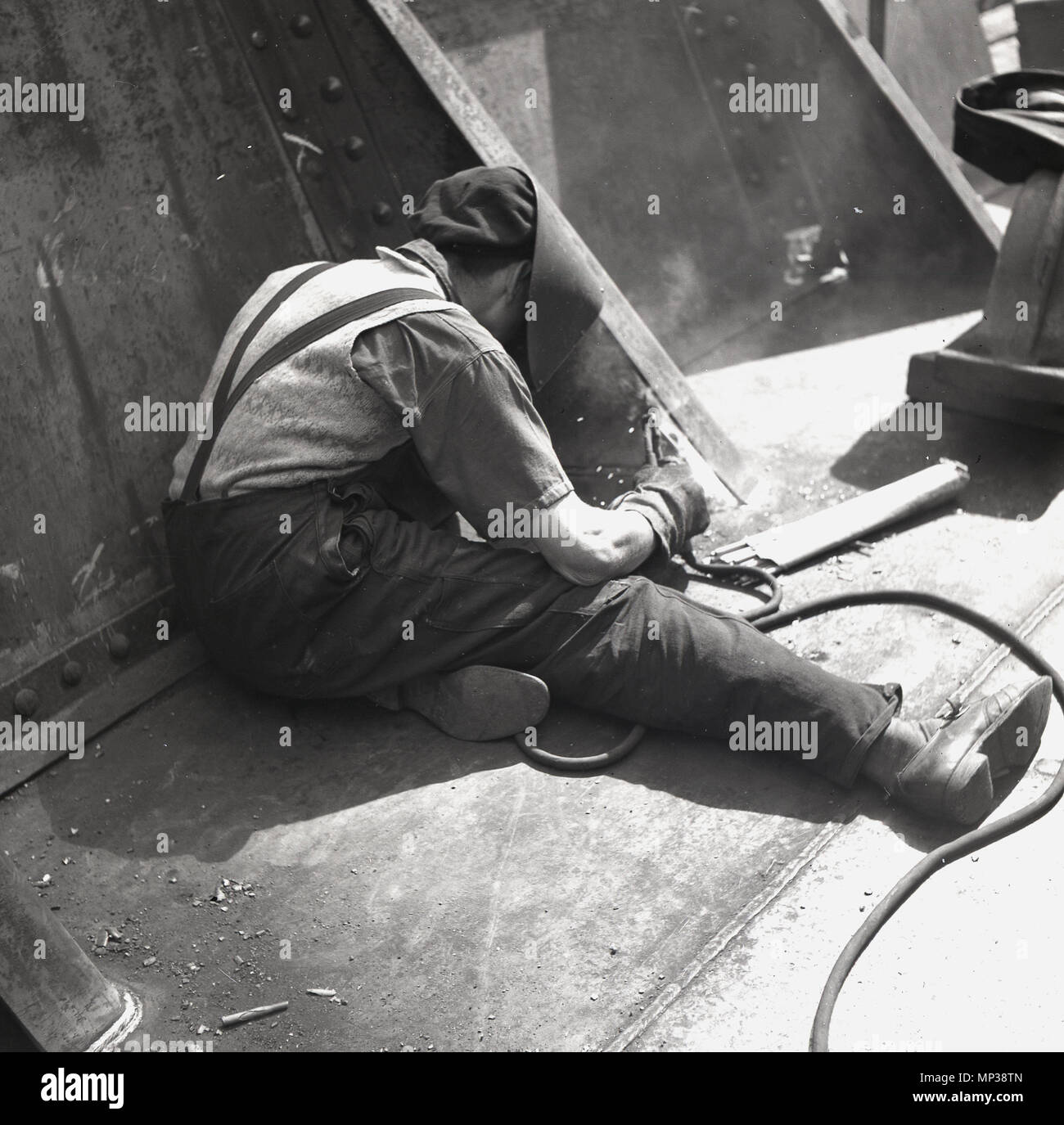 1950er Jahre, erwachsene männliche Arbeiter mit Helm oder ein Gesicht mit einem Schweißen Werkzeug Metallarbeiten Reparaturen auf eines Schiffsrumpfes, Belfast, Nordirland zu tun. Stockfoto